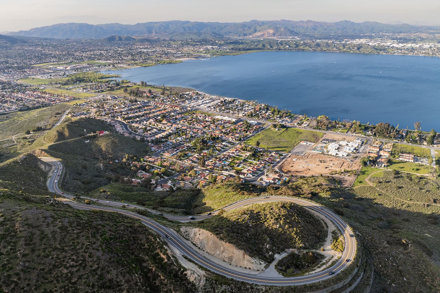 Aerial view of a winding road leading to a residential area by a large lake, surrounded by hills and distant mountains under a clear sky.