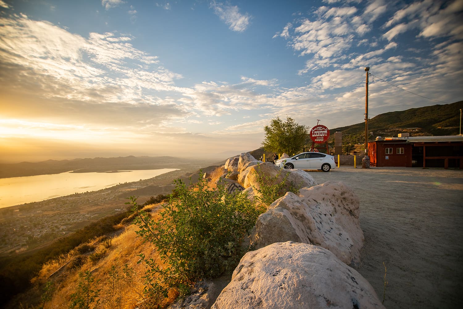 Scenic view of a sunset over a lake with a parked car and a small building near rocks in the foreground.