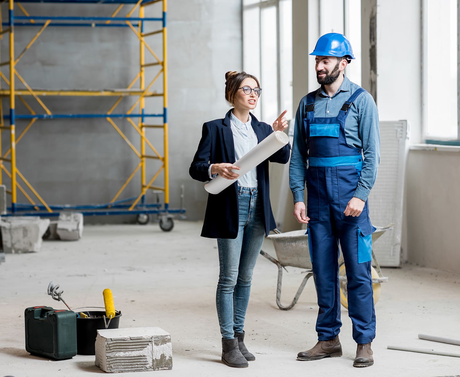 A woman holding a blueprint talks to a man in a hard hat and overalls at a construction site with scaffolding and tools in the background.