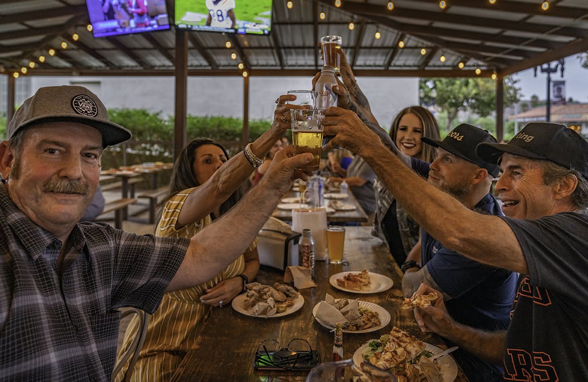 living-best-life Group of people toasting with drinks at a wooden table, plates of food in front of them, under string lights in an outdoor setting.