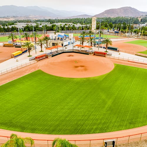 Aerial view of a baseball field with green grass and dirt infield, surrounded by palm trees and buildings in the background.