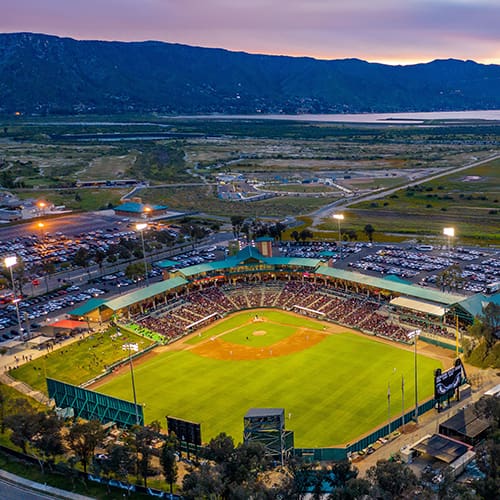Aerial view of a lit baseball stadium surrounded by parking lots and scenery with mountains in the background.