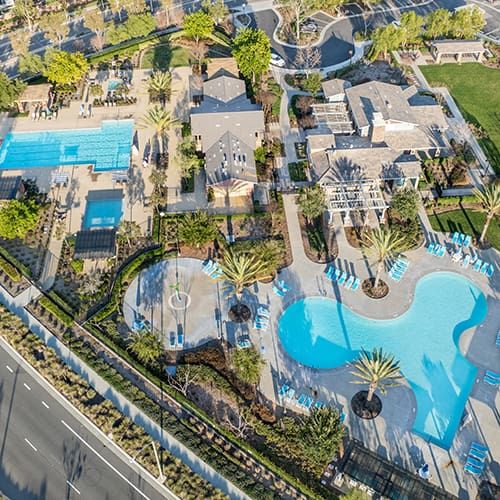 Aerial view of a residential area with two swimming pools, surrounding greenery, and nearby roads.