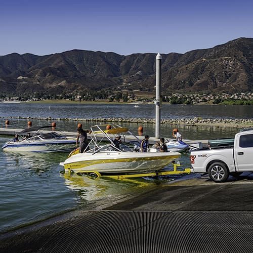 A truck is launching a yellow speedboat into a lake at a dock, with mountains in the background and another boat nearby.