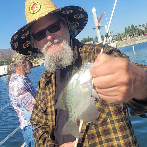 A man with a straw hat and sunglasses holds a small fish on a boat, with another person in the background and a body of water nearby.