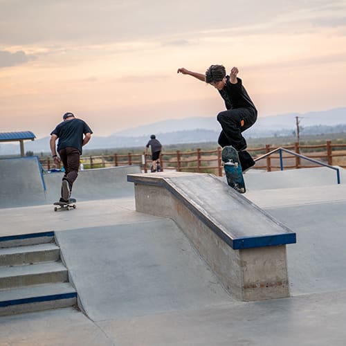 Skateboarders practice tricks at an outdoor skate park with ramps and rails during sunset.