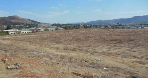 A vacant, dry dirt lot with scattered debris, bordered by buildings, a road, and mountains in the background under a clear blue sky.