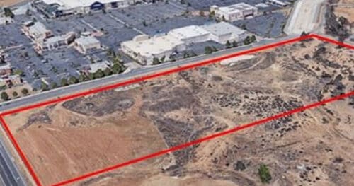 Aerial view of an undeveloped plot of land outlined in red, adjacent to a shopping center and paved roads.