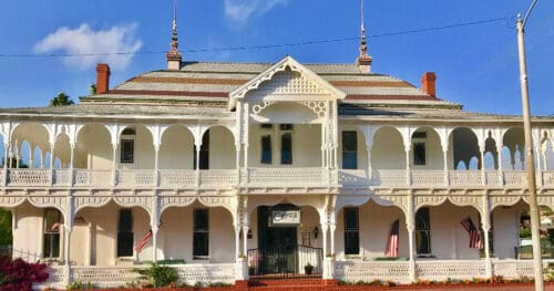 A large, historic two-story white building with ornate woodwork, a covered balcony, and three American flags displayed on the porch.