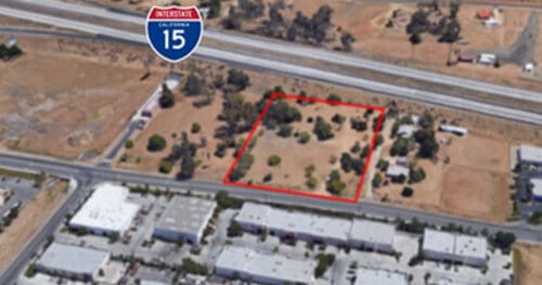 Aerial view of a fenced plot of land near industrial buildings with Interstate 15 and its road sign visible in the background.