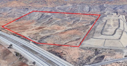 Aerial view of a hilly, undeveloped land area outlined in red, adjacent to highways and partially graded land.