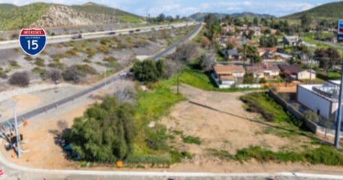 A vacant lot next to a road and freeway, with houses and hills in the background; an Interstate 15 highway sign is visible.