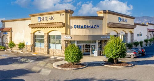 Exterior view of a Rite Aid pharmacy building with a parking lot, trees, and clear sky in the background.