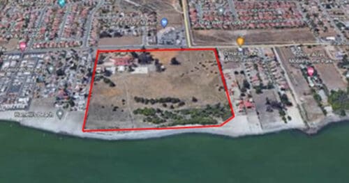 Aerial view of a coastal area with a section outlined in red, showing a mix of open land, trees, buildings, and adjacent residential neighborhoods.