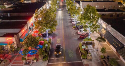 A car drives down a well-lit, tree-lined street with outdoor dining areas and parked cars on both sides at night.