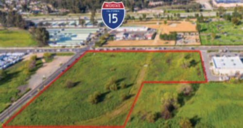 Aerial view of a grassy vacant lot outlined in red near a road, with Interstate 15 sign and commercial buildings in the background.