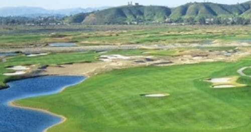 A golf course with green fairways, sand bunkers, and water hazards, set against a backdrop of hills and distant buildings.