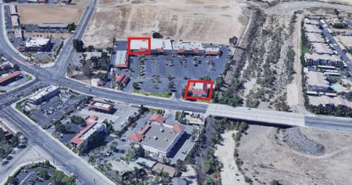 Aerial view of a commercial area with multiple buildings, two of which are outlined in red, next to a main road and a dry open field.