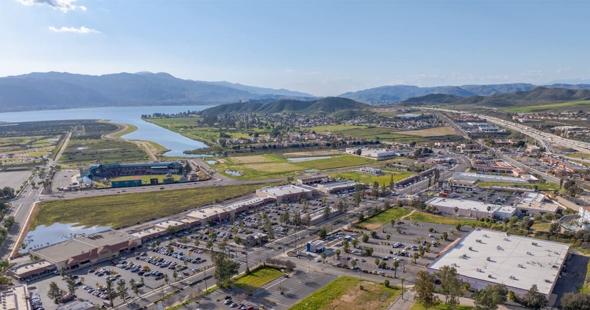 Aerial view of a suburban area with shopping centers, roads, parked cars, green fields, and a lake—set against mountains under a clear sky—showcasing growth as highlighted in the State of the City 2025 Wins.