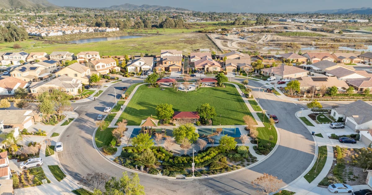 Aerial view of a suburban neighborhood near Summerly Golf Course in Lake Elsinore, with houses encircling a rectangular green park featuring trees, walkways, and a small playground—a perfect spot for families to enjoy a new chapter.