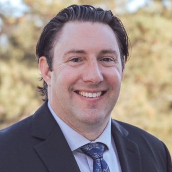 Man in a suit and tie smiling in an outdoor setting with blurred trees in the background.