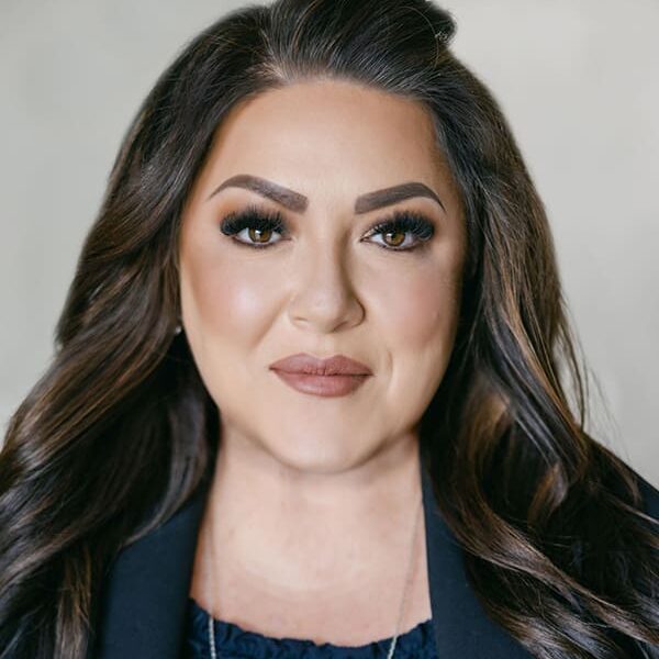 A woman with long brown hair wearing a dark blazer and navy top, looking straight at the camera against a neutral background.