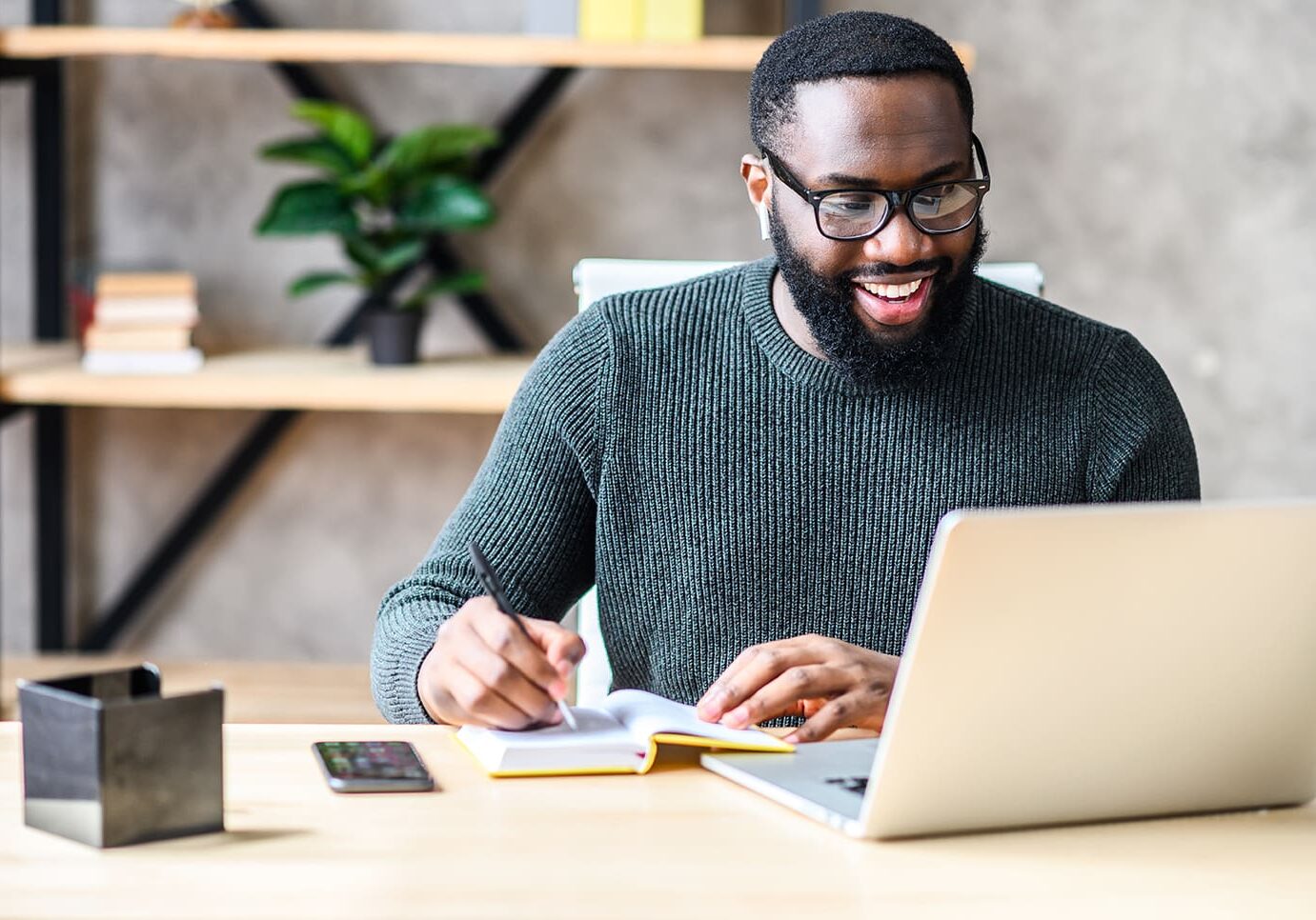 Man wearing glasses, smiling while working at a laptop, and taking notes at a desk.