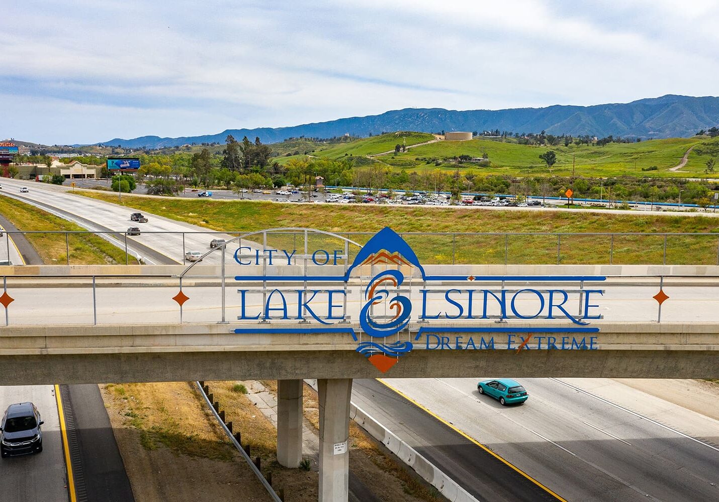 Bridge with City of Lake Elsinore, Dream Extreme signage over a highway with cars, surrounded by grassy landscape and distant hills.