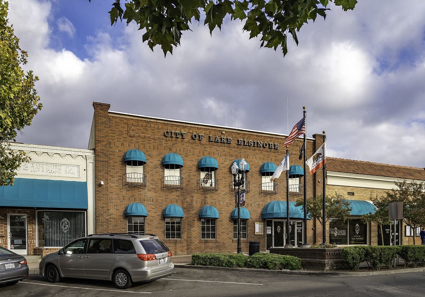 Street view of City of Lake Elsinore building, featuring red brick, blue awnings, and flagpoles. Cars are parked along the street, and a tree partially frames the top of the image.