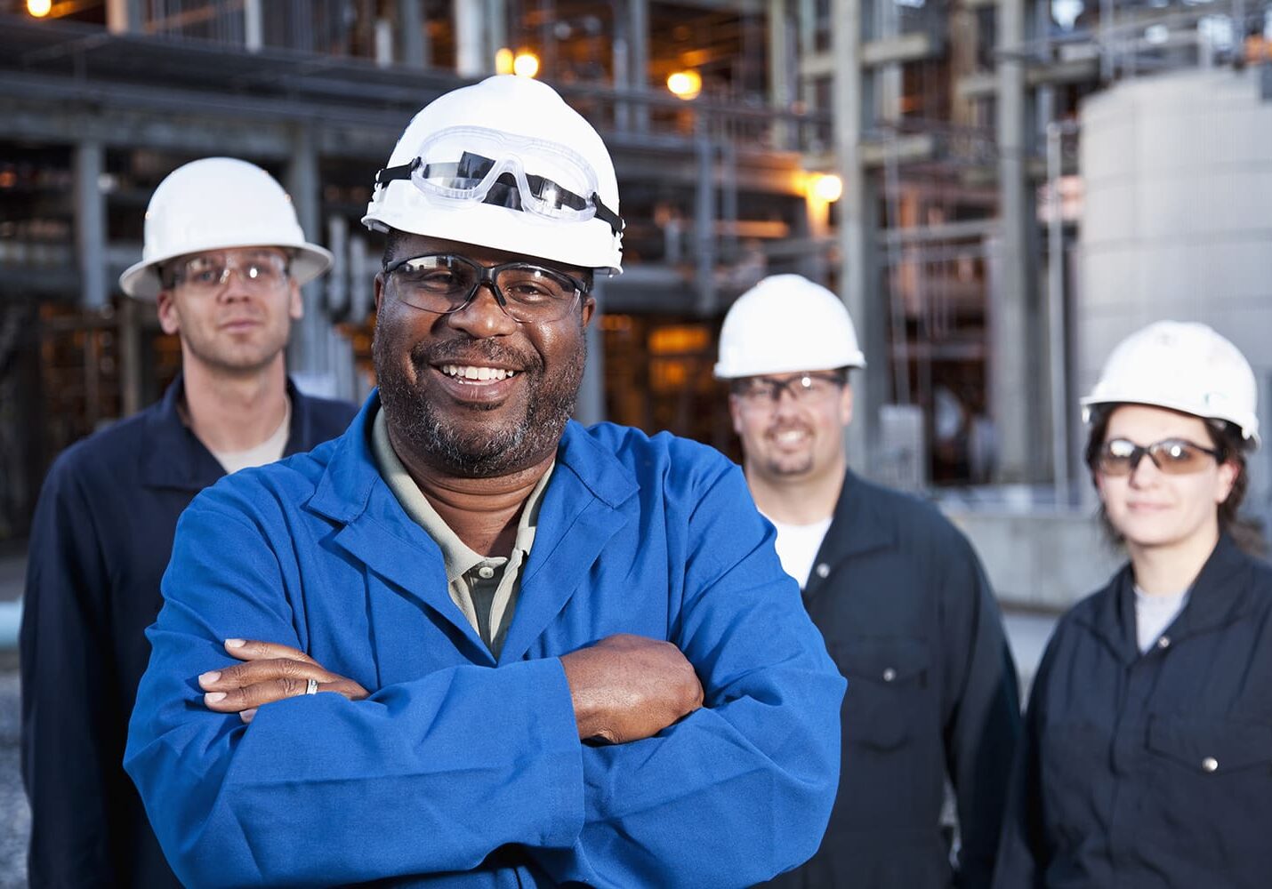 Four industrial workers in safety gear and hard hats stand in front of a factory background. One man in a blue jacket is in the foreground, smiling with arms crossed.