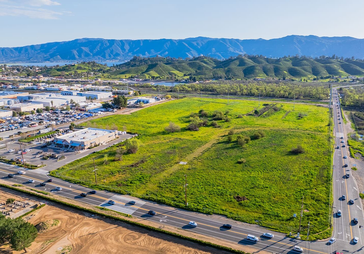 Aerial view of a green field near a busy road, surrounded by commercial buildings and distant hills under a clear sky.